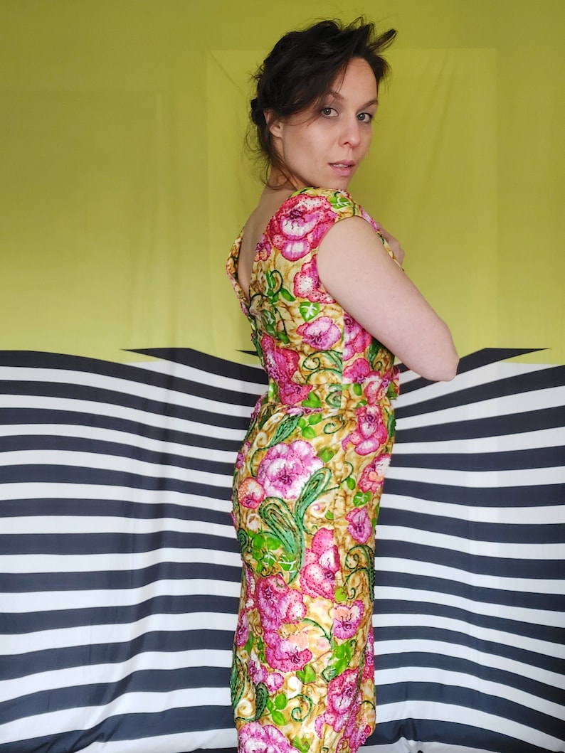 Op de afbeelding: Een vrouw draagt een roze en groene bloemenprintjurk met een ronde hals en korte mouwen. De jurk heeft een aansluitende pasvorm en is gemaakt van een glanzende stof.