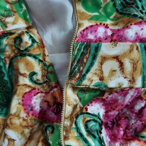 Op de afbeelding: Close-up van een rits op een kledingstuk met een kleurrijke bloemenprint. De stof is versierd met groene, roze en witte kralen.