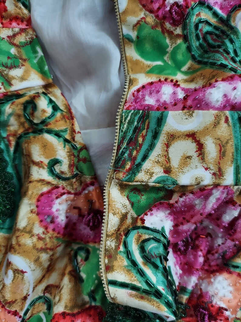 Op de afbeelding: Close-up van een rits op een kledingstuk met een kleurrijke bloemenprint. De stof is versierd met groene, roze en witte kralen.