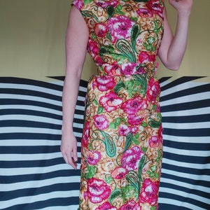 Op de afbeelding: Een vintage bloemenjurk met een roze, groene en gouden patroon. De jurk heeft een getailleerd bovenstuk en een volle rok. Het heeft een riem in de taille.