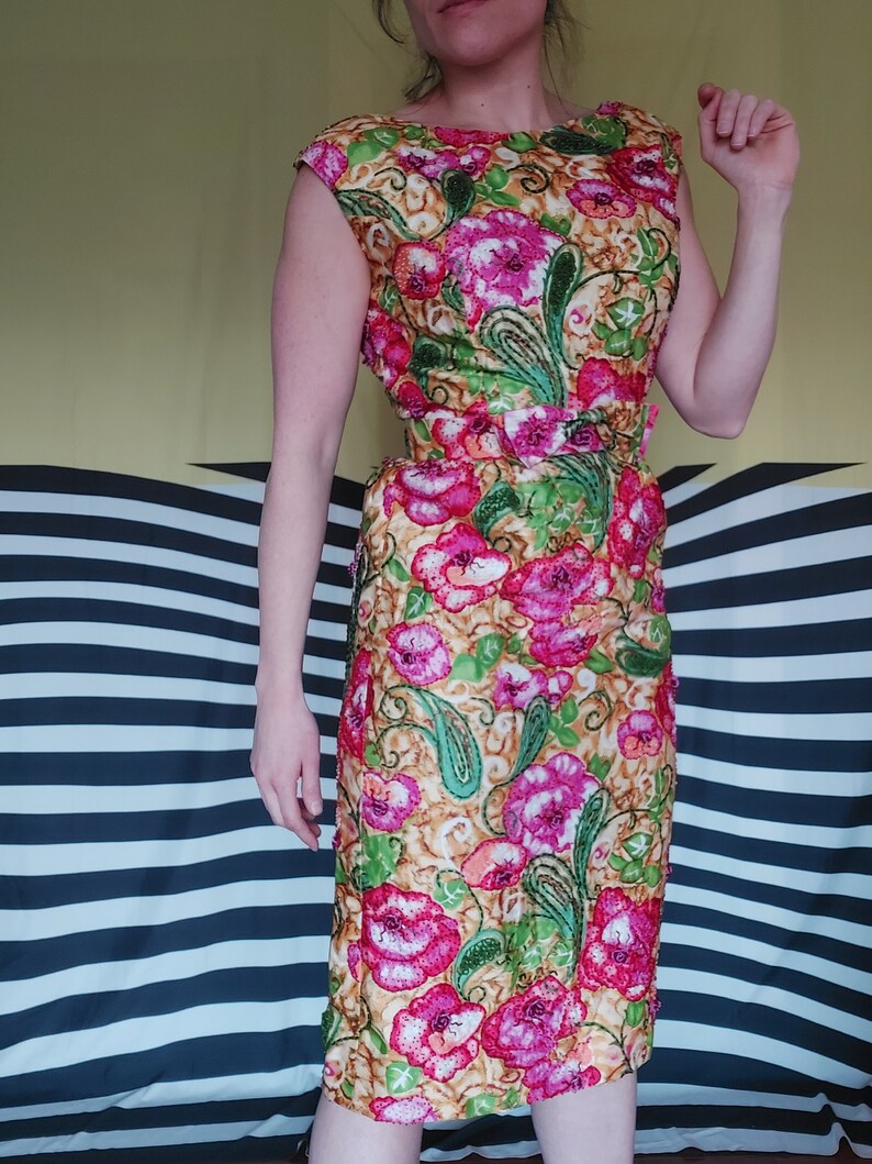 Op de afbeelding: Een vintage bloemenjurk met een roze, groene en gouden patroon. De jurk heeft een getailleerd bovenstuk en een volle rok. Het heeft een riem in de taille.
