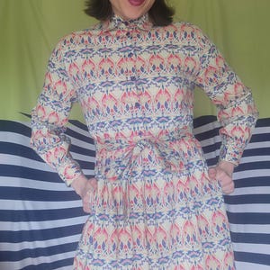 May include: A long-sleeved button-down dress featuring a repeating pattern of pink, blue, and cream floral motifs. The dress includes a collared neckline, a front button placket, and a fabric tie at the waist, creating a structured silhouette.