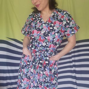 May include: A short-sleeved dress featuring a dark navy blue base with a vibrant floral pattern in shades of red, pink, light blue, and white. The dress includes a V-neckline, a tie-waist belt, and two visible front pockets at the hip area.