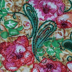 Op de afbeelding: Een close-up van een stof met een bloemenpatroon. De bloemen zijn roze, rood en wit met groene bladeren. De stof is versierd met kralen.