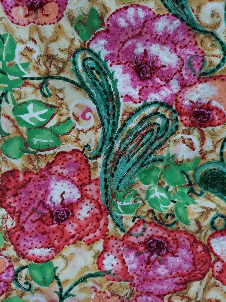 Op de afbeelding: Een close-up van een stof met een bloemenpatroon. De bloemen zijn roze, rood en wit met groene bladeren. De stof is versierd met kralen.
