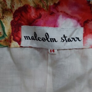Op de afbeelding: Een wit label met zwarte tekst die "malcolm starr" luidt en een wit label met rode tekst die "14" luidt. Het label is bevestigd aan een witte stof met een bloemenpatroon.