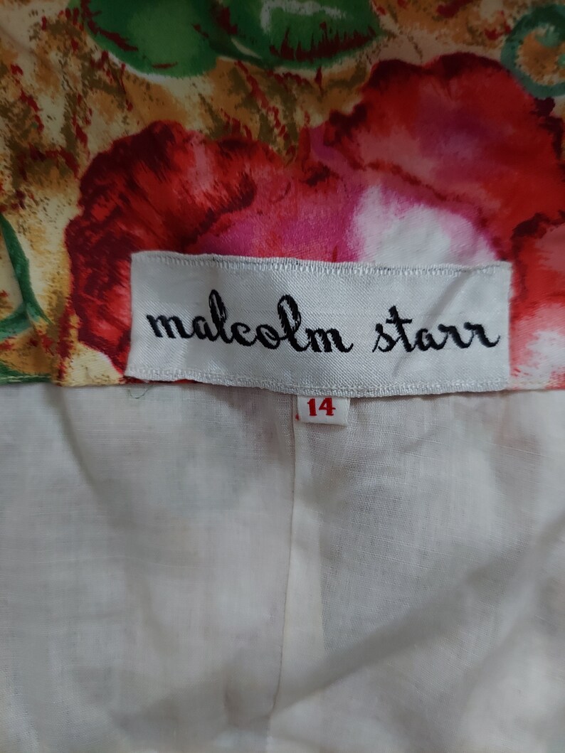 Op de afbeelding: Een wit label met zwarte tekst die "malcolm starr" luidt en een wit label met rode tekst die "14" luidt. Het label is bevestigd aan een witte stof met een bloemenpatroon.