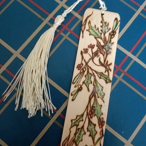 May include: A wooden bookmark with a cream tassel. The bookmark has a pyrography design of holly leaves, berries, and mistletoe, with green and red accents. The background is a blue plaid pattern.