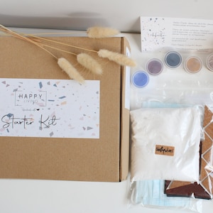 Könnte beinhalten: Ein Bastelset zum Herstellen von Terrazzo-Untersetzern. Das Set enthält eine braune Box mit der Aufschrift "Happy Living Starter Kit" auf der Vorderseite, einen Beutel mit weißem Pulver mit der Aufschrift "GipsPulver", sechs kleine Gläser mit farbigem Harz, ein transparentes Tablett aus Kunststoff, einen transparenten Kreis aus Kunststoff und einen Holzrührstab.