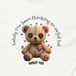 Op de afbeelding: Een wit t-shirt met een afbeelding van een beige teddybeer die aan elkaar is genaaid met diverse gekleurde spelden in het lichaam. De beer heeft knoopogen en een paarse hartvormige patch. De tekst luidt: Lately I've been thinking an awful lot ABOUT YOU.