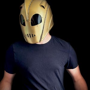 May include: A gold-colored helmet with a pointed top and riveted details. The helmet has eye openings with dark lenses and a mouth vent. The person is wearing a black t-shirt.