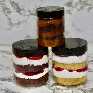 Könnte beinhalten: Drei Glasbehälter mit schwarzen Deckeln, gefüllt mit geschichteten Desserts. Die Behälter sind übereinander gestapelt. Die Desserts beinhalten Schokoladenkuchen, Vanillekuchen, Schlagsahne und Fruchtaufstrich.