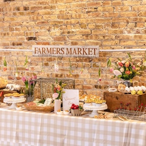 May include: A farmers market display with a sign that reads "FARMERS MARKET". The table is covered with a tan and white checkered tablecloth. The display includes flowers, baked goods, and fresh produce. The background is a brick wall.