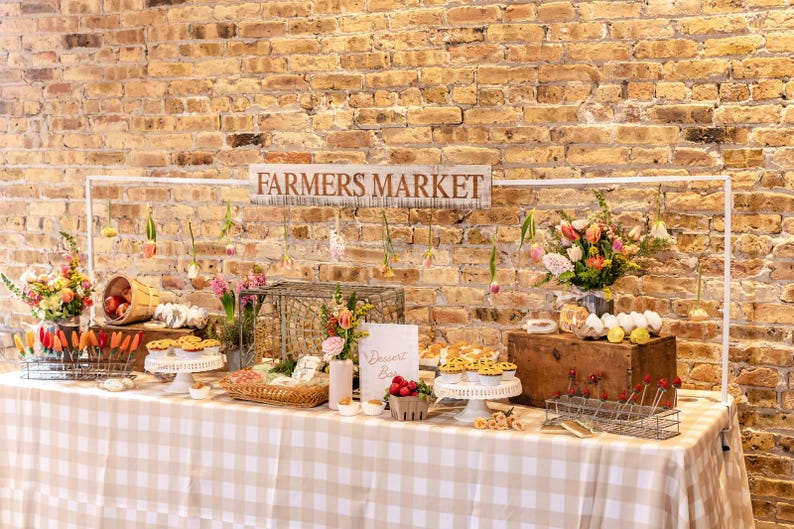 May include: A farmers market display with a sign that reads "FARMERS MARKET". The table is covered with a tan and white checkered tablecloth. The display includes flowers, baked goods, and fresh produce. The background is a brick wall.