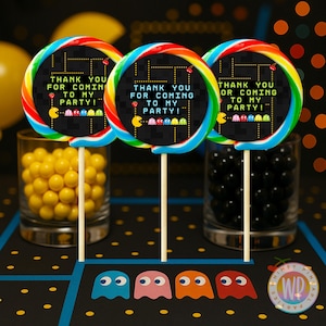 May include: Three rainbow-colored lollipops with a Pac-Man theme. Each lollipop has a black center with the text "THANK YOU FOR COMING TO MY PARTY!" and Pac-Man characters. The lollipops are on wooden sticks, with glasses of yellow and black candies in the background.