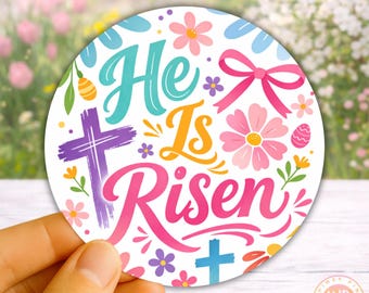 May include: A round, white Easter-themed design with the words "He Is Risen" in colorful script. The design includes crosses, flowers, a bow, and an Easter egg. The background is a blurred image of flowers.