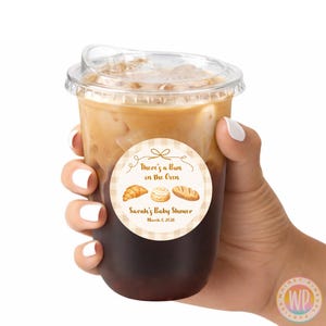 May include: A clear plastic cup filled with a layered coffee drink, held by a hand with white nail polish. A round sticker on the cup reads "There's a Bun in the Oven" with bakery illustrations, and "Sarah's Baby Shower, March 7, 2026."