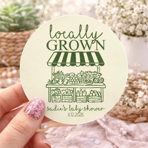 K&ouml;nnte beinhalten: Ein runder, cremefarbener Aufkleber mit einer dunkelgr&uuml;nen Strichzeichnung eines Marktstandes mit Produkten. Der Text lautet "locally GROWN" oben und "sadie's baby shower 6.12.2026" unten in einer dunkelgr&uuml;nen Schreib- und Sans-Serif-Schrift.