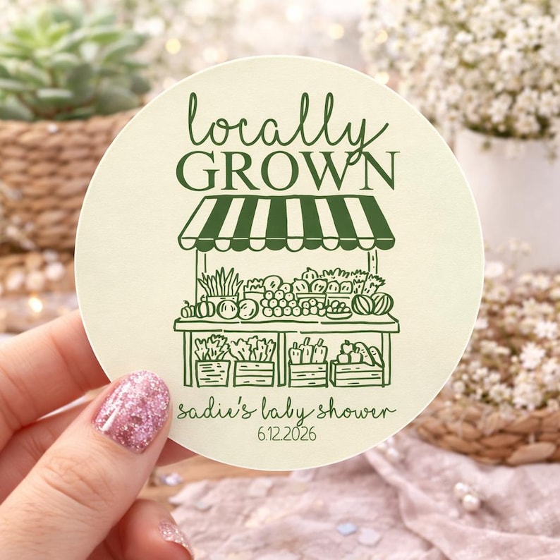 K&ouml;nnte beinhalten: Ein runder, cremefarbener Aufkleber mit einer dunkelgr&uuml;nen Strichzeichnung eines Marktstandes mit Produkten. Der Text lautet "locally GROWN" oben und "sadie's baby shower 6.12.2026" unten in einer dunkelgr&uuml;nen Schreib- und Sans-Serif-Schrift.