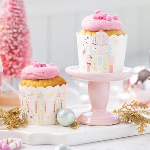 May include: A festive scene featuring cupcakes with pink frosting and decorative nutcracker figurines. The cupcakes are in patterned wrappers and sit on a pink cake stand. Additional ornaments and a pink Christmas tree complete the holiday theme.