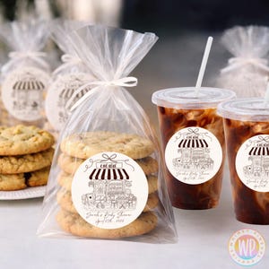 May include: A clear cellophane bag tied with a white ribbon contains six cookies with a sticker that says "CAFE BÉBÉ" and "Sarah's Baby Shower April 25th 2026". Two iced coffee drinks with straws are next to the bag.