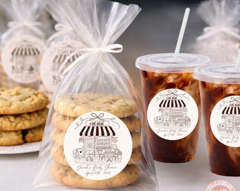 May include: A clear cellophane bag tied with a white ribbon contains six cookies with a sticker that says "CAFE BÉBÉ" and "Sarah's Baby Shower April 25th 2026". Two iced coffee drinks with straws are next to the bag.