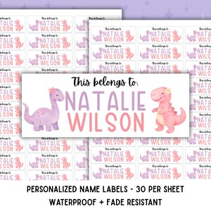 Peut inclure: Une feuille d'étiquettes nominatives rectangulaires personnalisées avec le texte This belongs to: NATALIE WILSON en violet et rose. Chaque étiquette comprend un dinosaure violet à gauche et un dinosaure rose à droite, portant des fleurs.