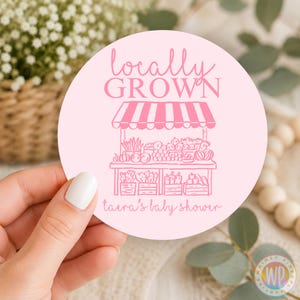 K&ouml;nnte beinhalten: Ein runder, hellrosa Aufkleber mit einer dunkelrosa Strichzeichnung eines Marktstandes mit Obst- und Gem&uuml;sekisten. Der Text lautet locally GROWN und taera's baby shower in einer kursiven und blockartigen Schriftart auf der Oberfl&auml;che.