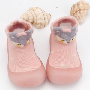 May include: A pair of pink baby booties with a grey animal face design. The booties are made of a soft, stretchy material and have a rubber sole.