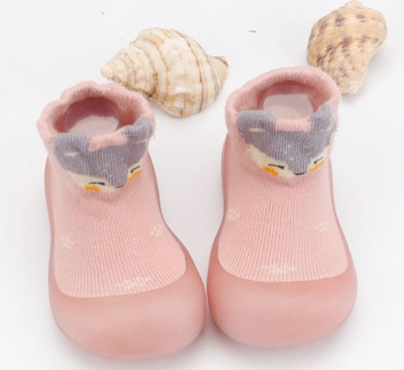 May include: A pair of pink baby booties with a grey animal face design. The booties are made of a soft, stretchy material and have a rubber sole.