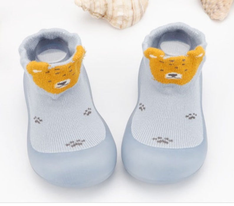 May include: A pair of light blue baby booties with a yellow bear face design on the front. The booties have a rubber sole and are made of soft, breathable fabric.