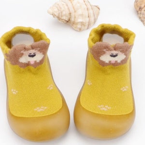May include: A pair of yellow baby booties with a brown dog face design on the front. The booties have a rubber sole.