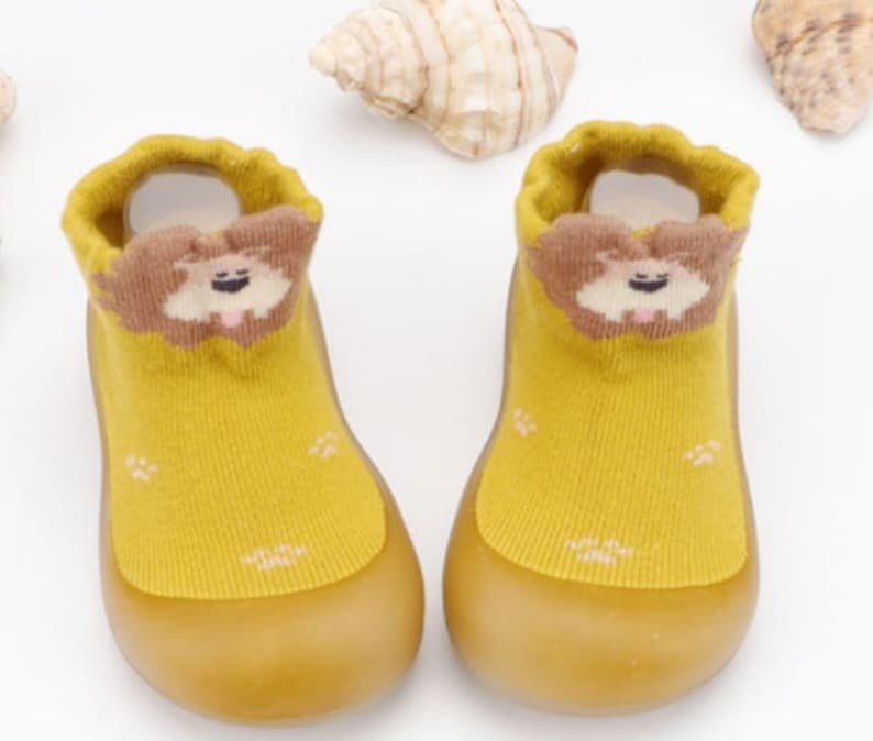 May include: A pair of yellow baby booties with a brown dog face design on the front. The booties have a rubber sole.