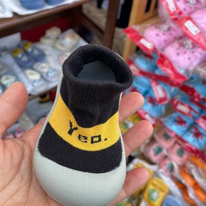 May include: A pair of grey baby booties with black and yellow trim. The booties have the word "Yeo" written in yellow on the black trim.