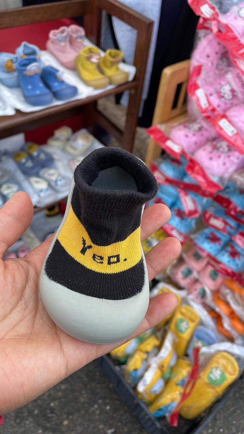 May include: A pair of grey baby booties with black and yellow trim. The booties have the word "Yeo" written in yellow on the black trim.