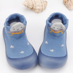 May include: A pair of blue baby booties with a white cloud and star design. The booties have a rubber sole and are made of soft, breathable fabric.