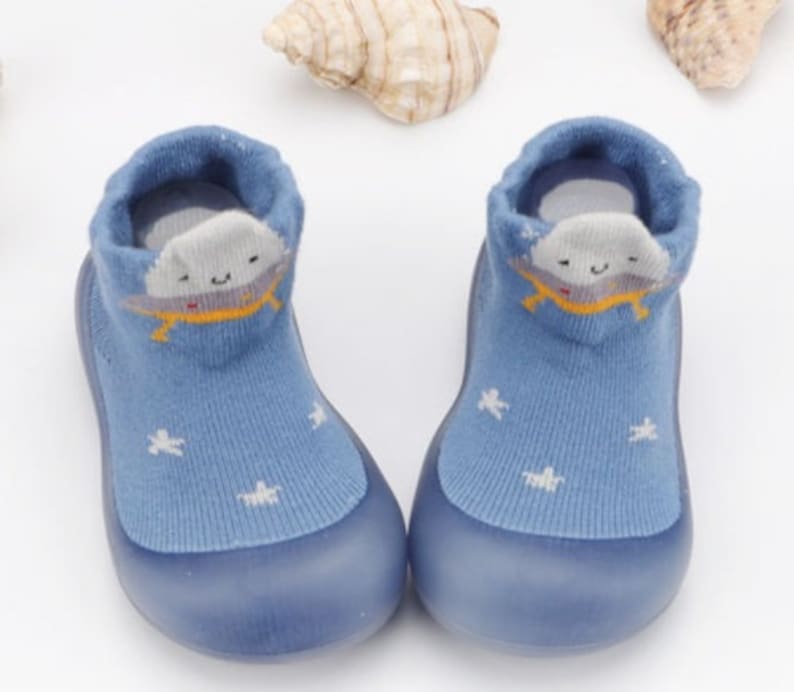 May include: A pair of blue baby booties with a white cloud and star design. The booties have a rubber sole and are made of soft, breathable fabric.