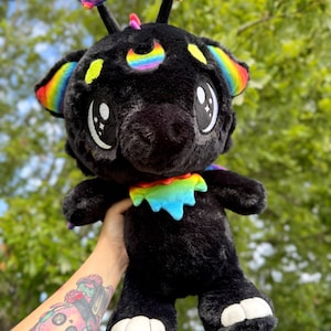 以下が含まれることがあります： 虹色のアクセントが付いた黒いぬいぐるみ。耳、三日月、星型のアンテナが含まれます。ぬいぐるみは大きく、表情豊かな目をしており、足には白いアクセントが付いています。