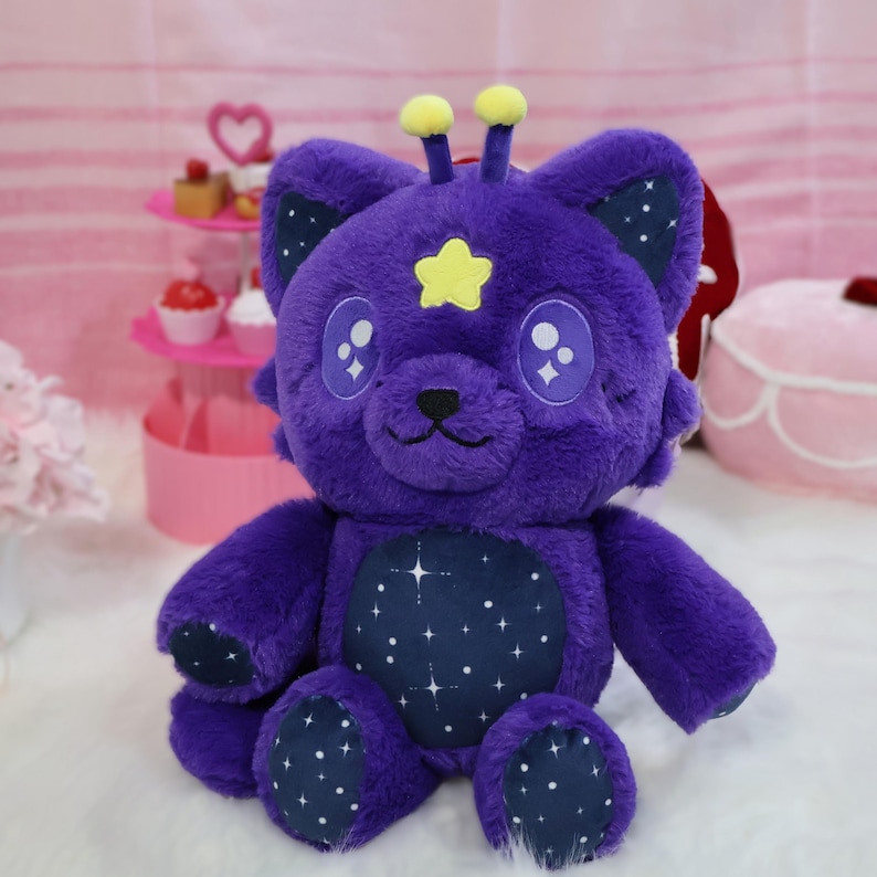 Op de afbeelding: Een paarse teddybeer met een stervormig gezicht en een sterrenbuik. De beer heeft gele antennes en sterrenaccenten op zijn oren en poten. De beer zit op een wit oppervlak.
