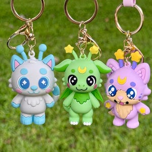 May include: Three colorful cartoon character keychains. The keychains feature a light blue and white cat-like creature, a green dragon-like creature, and a purple cat-like creature. Each keychain has a gold-colored ring and clasp.