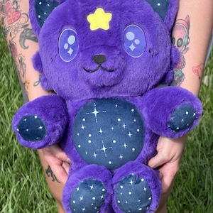 Op de afbeelding: Een paars, katachtig pluche speelgoed met een gele ster op het voorhoofd en een sterrenbuik. Het speelgoed heeft grote, expressieve ogen en kleine gele antennes. De poten en oren hebben een donkerblauwe stof met witte sterrenpatronen.