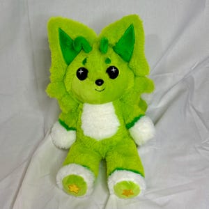 May include: A plush toy in shades of green, featuring a stylized animal design. The toy has large, pointed ears, a white chest, and white accents on the paws. The eyes are black with white star-shaped highlights, and the feet have yellow star details.