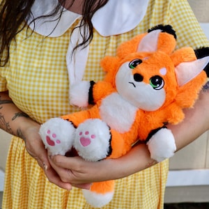 May include: An orange plush fox toy with white accents, black-tipped ears, and pink paw pads. The fox has large green eyes and a sad expression. The plush toy is being held in front of a yellow gingham dress.