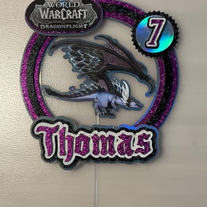 May include: A circular cake topper featuring a World of Warcraft Dragonflight logo, a blue and black dragon illustration, the number 7 in a holographic circle, and the name Thomas in purple glittery font, all set against a black and purple frame.