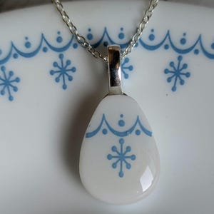 May include: A white pendant necklace with a silver chain. The pendant has a teardrop shape and features a blue snowflake design. The pendant is set against a white background with blue snowflake patterns.