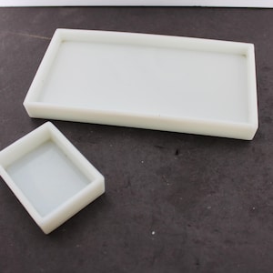 May include: Two rectangular white trays with raised edges. The larger tray is elongated and positioned horizontally, while the smaller tray is compact and placed at an angle. Both items feature a smooth, solid white finish and a simple, open design.