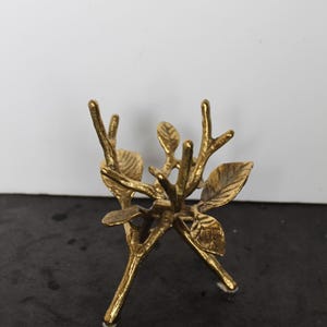 May include: A gold-colored, decorative metal branch with leaves. The sculpture has three legs and is designed to hold small items. The leaves and branches are detailed, and the overall design is nature-inspired.