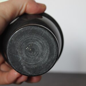 May include: A dark gray, cylindrical object with a rounded base. The base has concentric circular grooves. The object appears to be made of a solid, possibly stone or ceramic material. The object is held in a person's hand.
