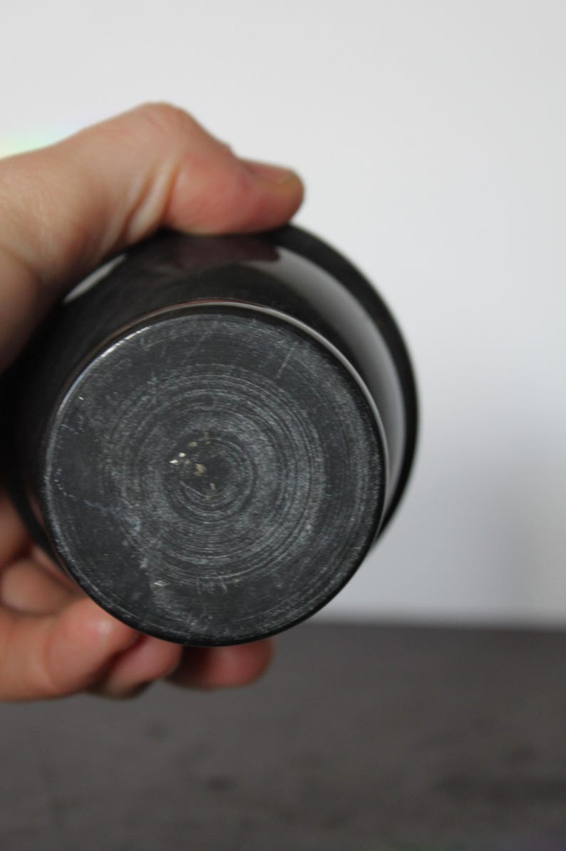 May include: A dark gray, cylindrical object with a rounded base. The base has concentric circular grooves. The object appears to be made of a solid, possibly stone or ceramic material. The object is held in a person's hand.