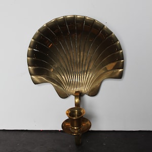 May include: A brass seashell-shaped wall sconce with a candle holder. The sconce has a fan-like design with vertical lines and a curved edge. The candle holder is round and has a small base. The overall color is gold.